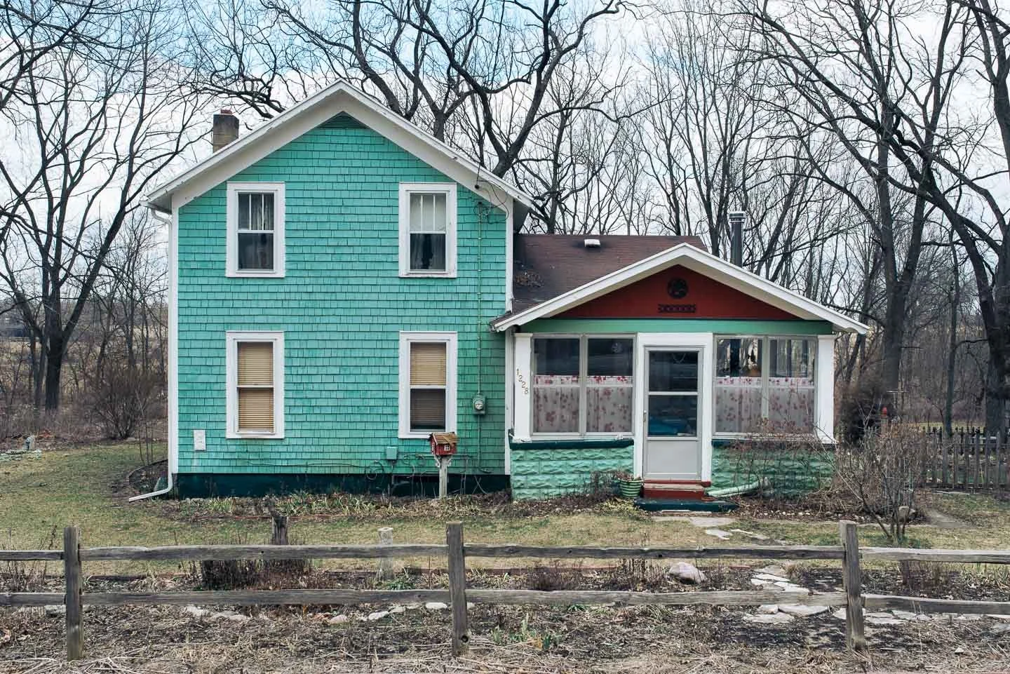 A two-story house with turquoise siding and white trim, attached to a one-story enclosed porch with red and green accents, surrounded by leafless trees and a wooden fence.