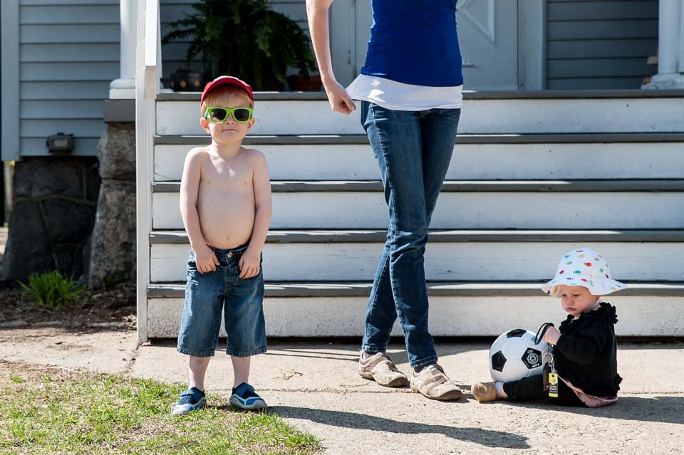 A shirtless boy wearing green sunglasses and denim shorts standing outside next to a seated baby girl with a hat, holding a soccer ball.