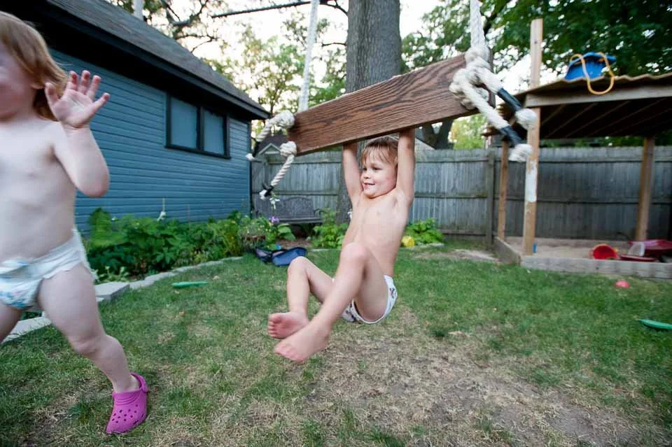Two young children playing in a backyard with a swing set; one child is sitting on the swing, holding onto the wooden plank, and smiling, while the other child is standing nearby. The backyard has a blue house, green plants, and a wooden fence, with 