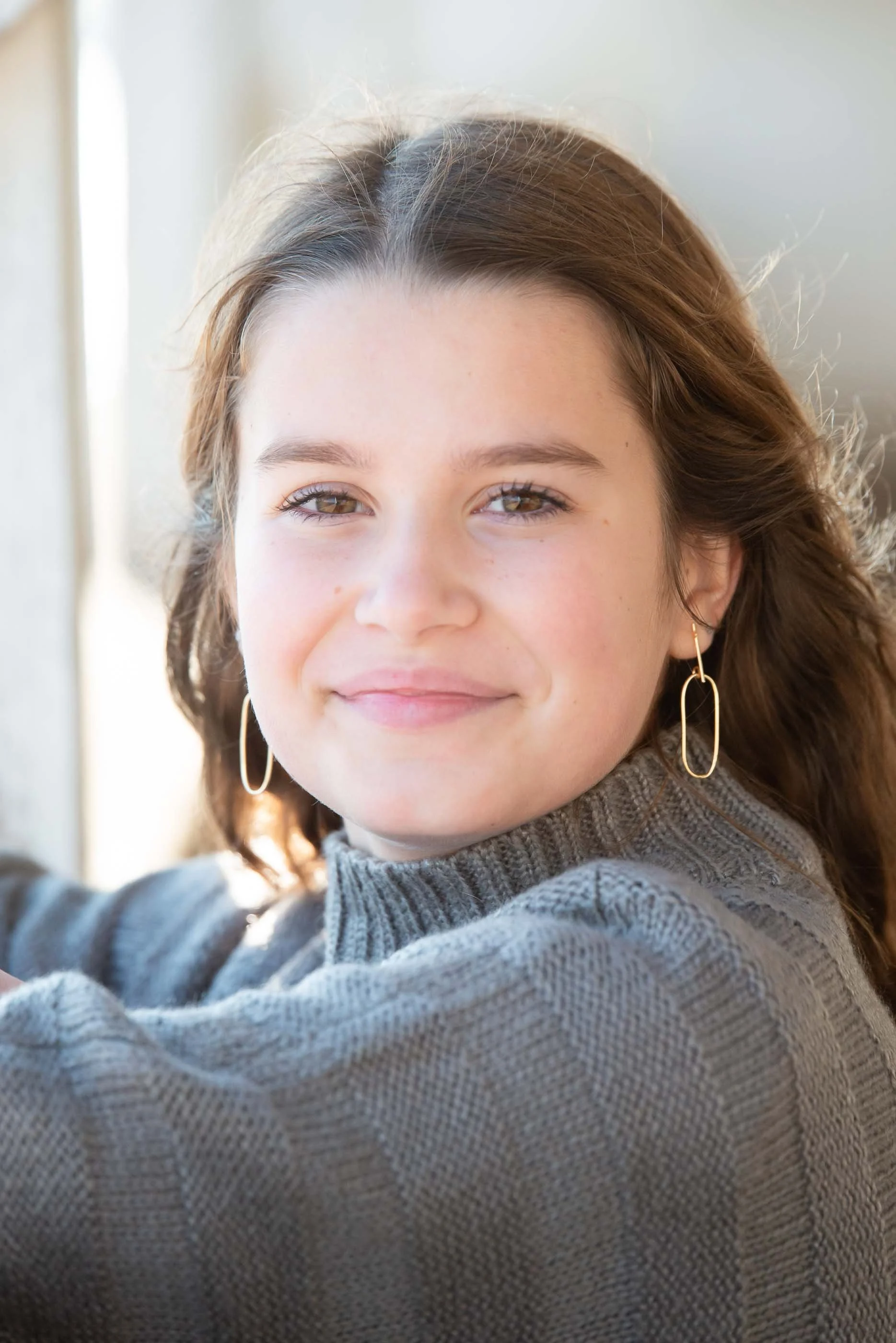 A young woman with brown hair and brown eyes smiling at the camera, wearing gold hoop earrings and a gray knitted sweater, indoors near a bright window.