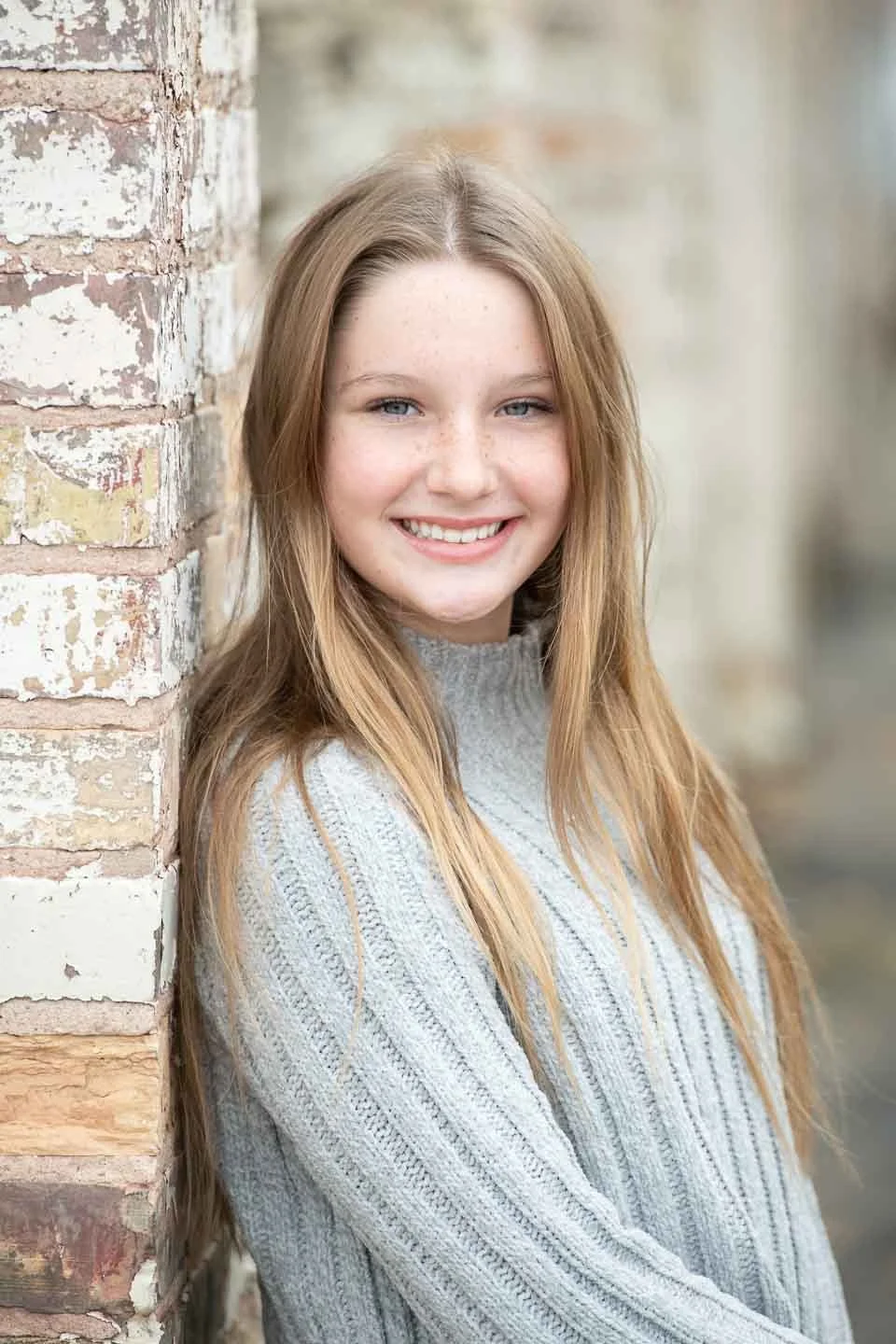 A young woman with long, blonde hair smiling while leaning against a weathered brick wall outdoors.
