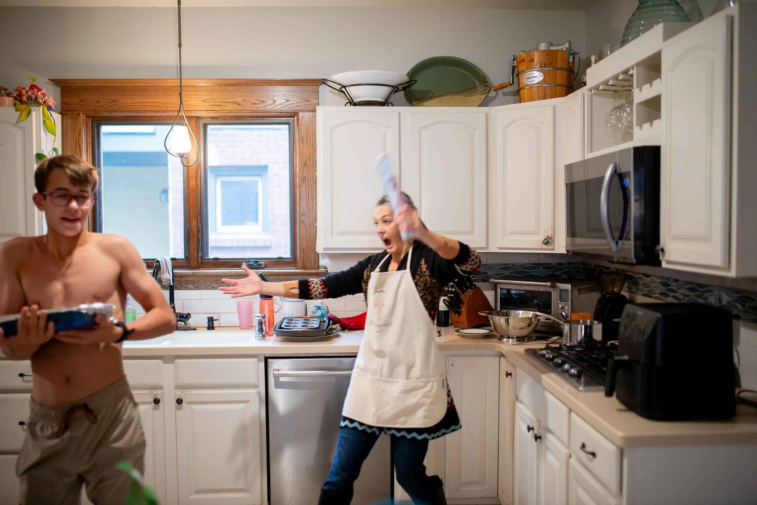 A woman in a kitchen is throwing a roll of paper towels at a shirtless boy holding a package of food. The woman appears angry or upset, while the boy looks surprised. The kitchen has white cabinets, a microwave, and various kitchen appliances.