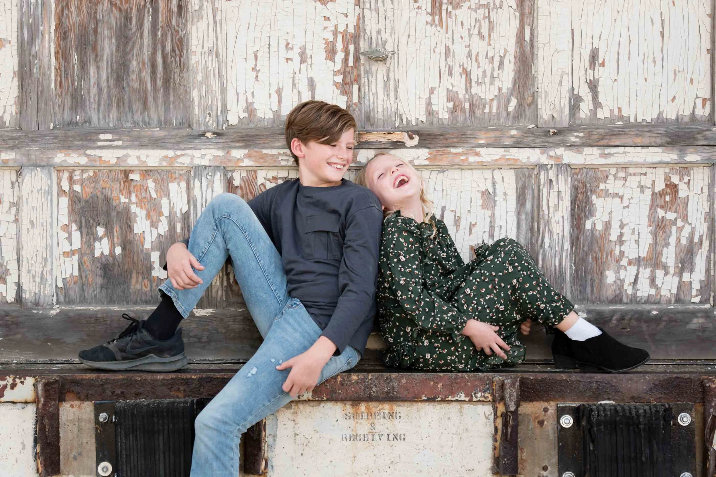 Two children, a boy and a girl, sitting on a weathered wooden surface outdoors, laughing and enjoying each other's company.