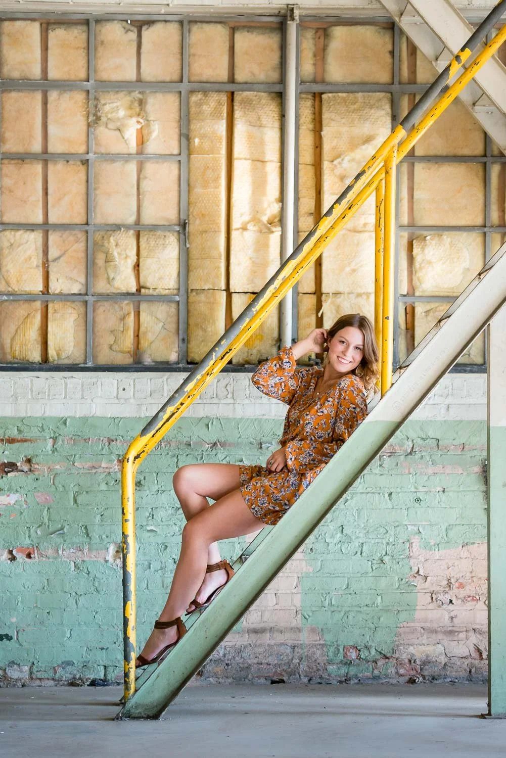 A woman sitting on a staircase with a colorful, partially exposed brick wall behind her. She is smiling, wearing a floral dress, and adjusting her hair.
