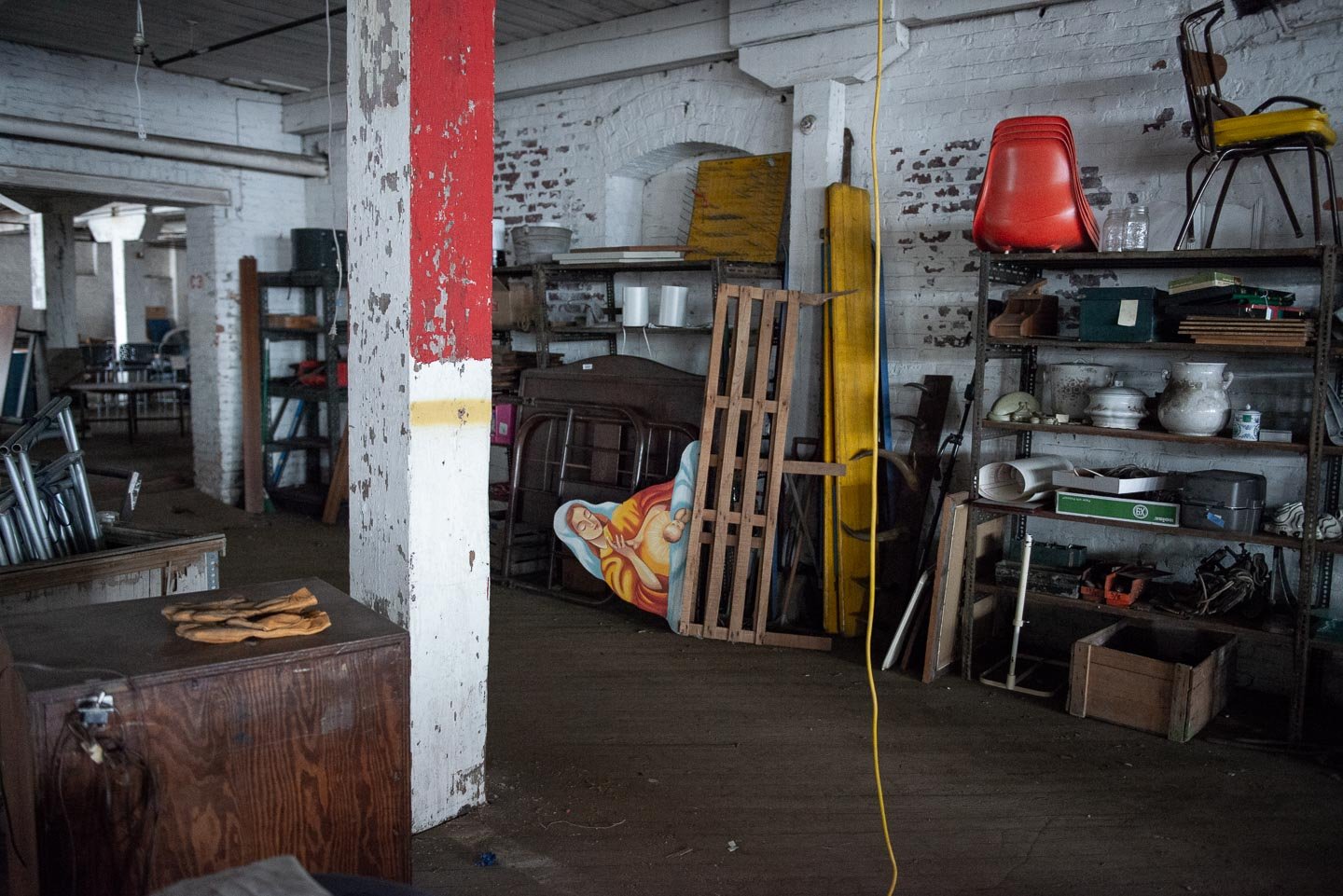 Inside an old, cluttered storage space or warehouse with white painted brick walls, a red and white painted pillar, shelves filled with various items including dishes, chairs, a yellow surfboard, and a cardboard cutout of a person. The floor is woode