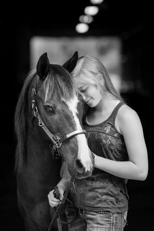 A young woman with blonde hair gently resting her forehead against a horse's face, both eyes closed, inside a stable or indoors.