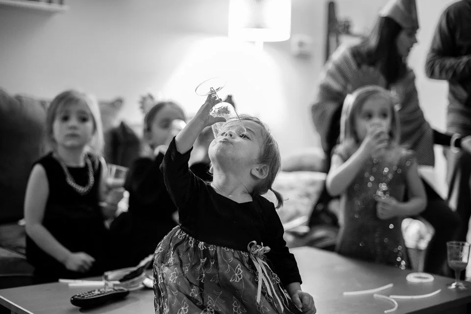 A young girl holding a toy helicopter up to her nose, with other children and adults in the background at a party.