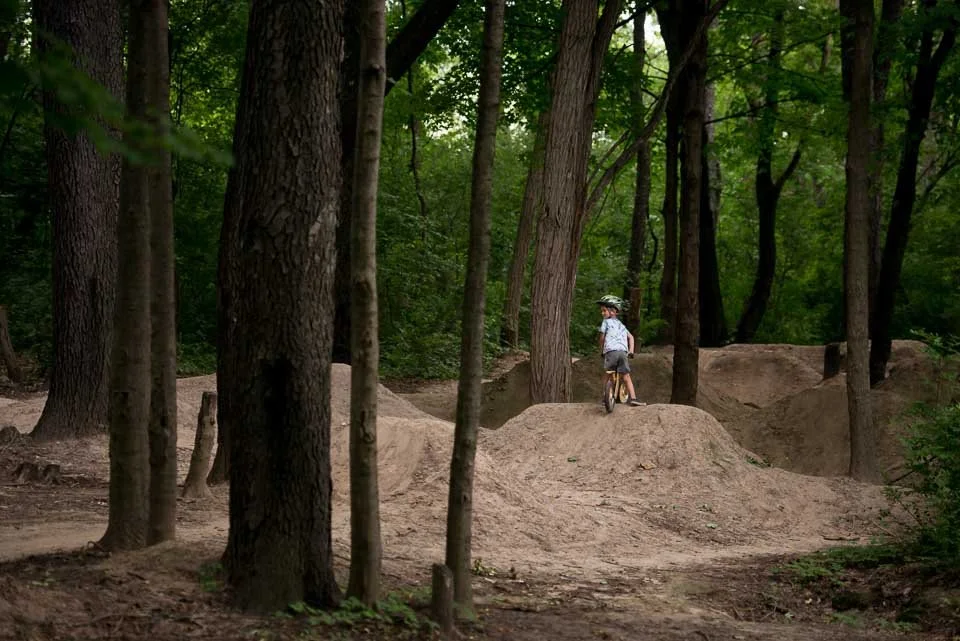 A child riding a bike on dirt jumps in a wooded area with tall trees and green foliage.