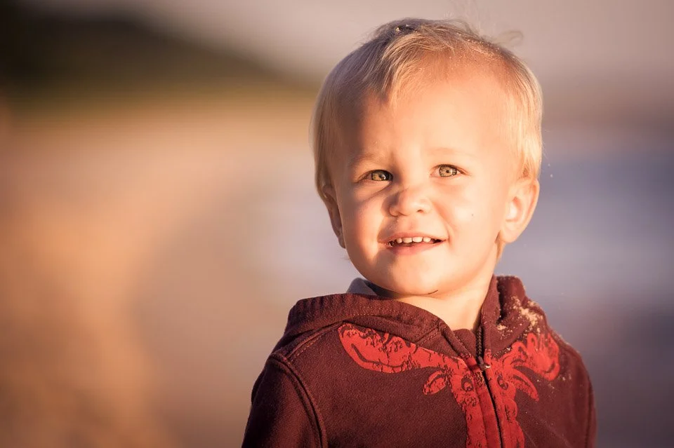 A young boy with blonde hair and light skin standing outdoors near a body of water, smiling and looking to the side, wearing a dark red hoodie with a dragon design.