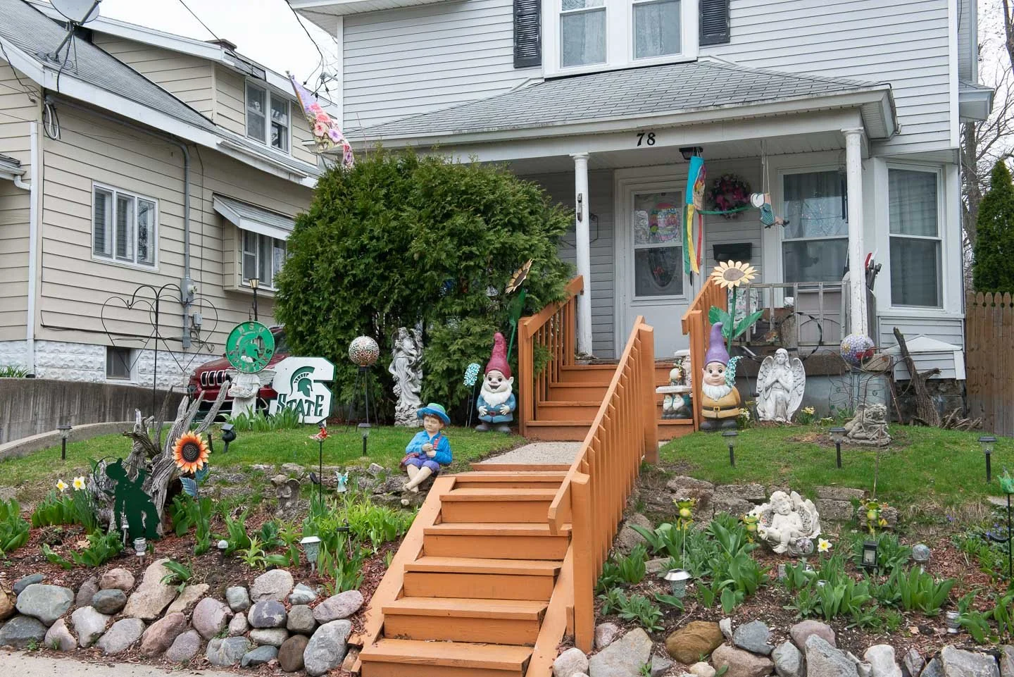 Front yard garden decorated with garden gnomes, stone angel statues, a sunflower decoration, and a Michigan State University sign. The house has a wooden staircase leading to the porch, with colorful wind chimes and flags hanging on the porch. A gard