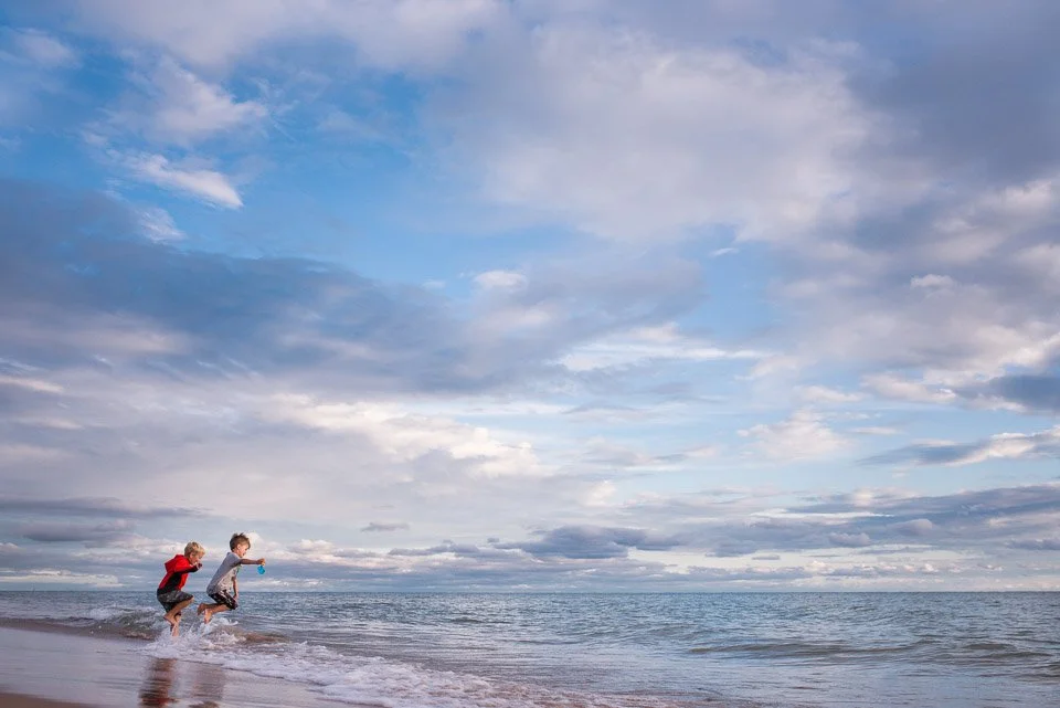 Two children playing and running in the shallow water at a beach during daytime with a partly cloudy sky.