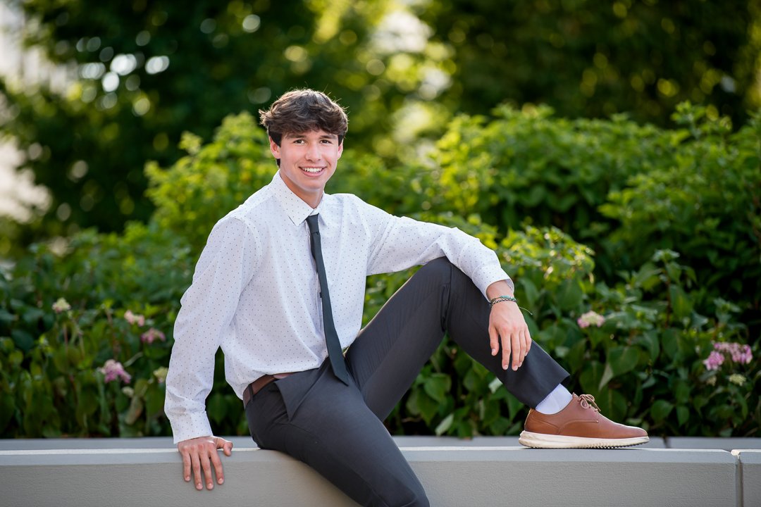 Senior portrait in a dress shirt and tie sitting on a ledge among greenery in downtown Grand Rapids