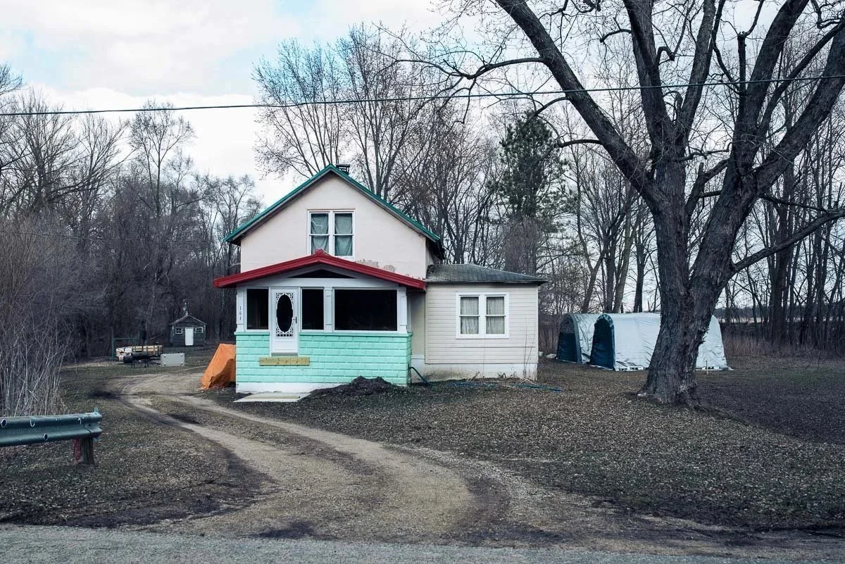 A white two-story house with a red roof and trim, featuring a covered front porch and a dirt driveway, surrounded by leafless trees with small structures visible in the background.