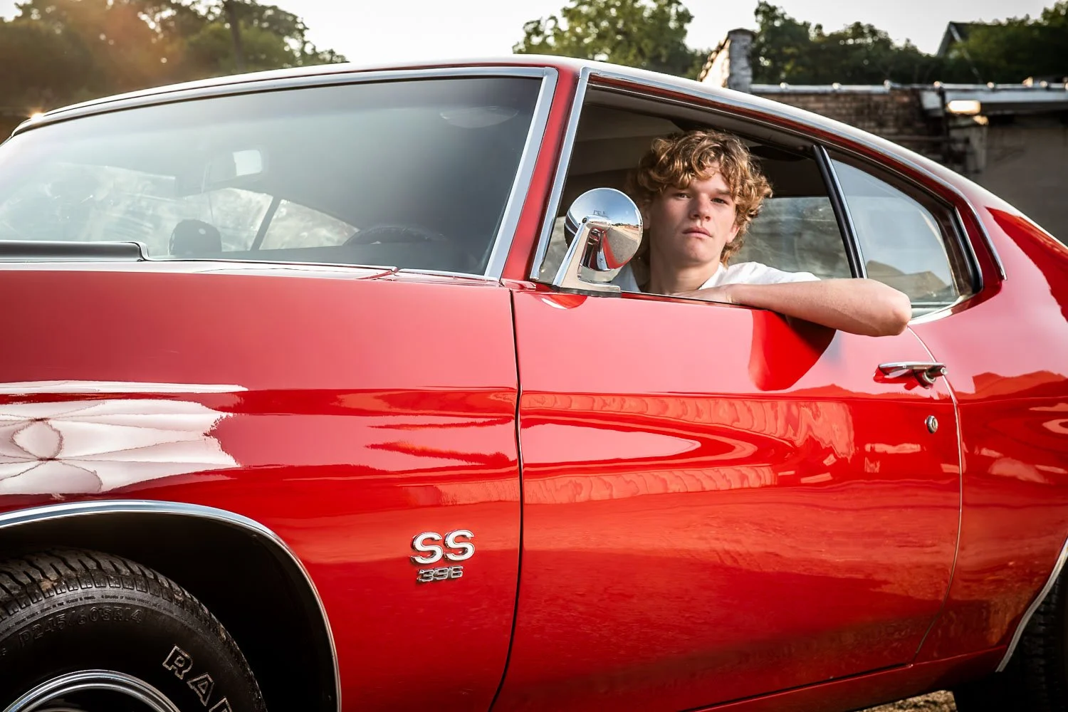 A young man with curly hair sitting in the driver's seat of a red vintage Chevrolet car, looking out the open window with a serious expression. The car has an 'SS 396' badge on the side and is parked outdoors with a background of trees and a building