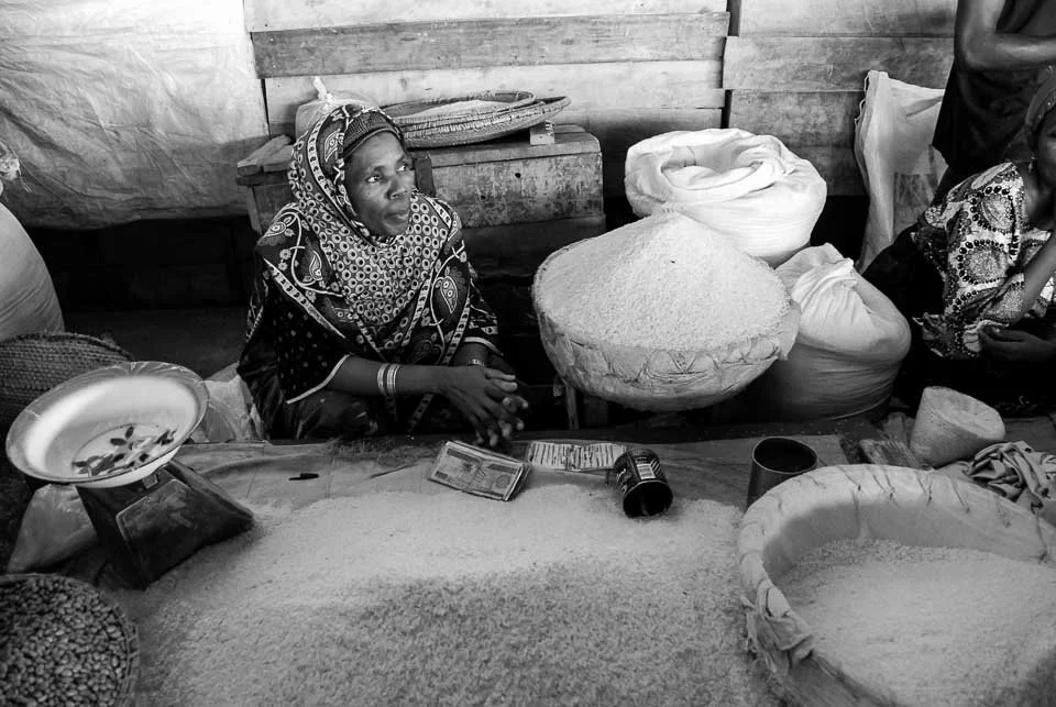 A woman wearing a patterned headscarf and traditional clothing sits behind a table with various sacks of grains or flour, surrounded by weighed items, a scale, and a phone. The setting appears to be a marketplace or a storage area.