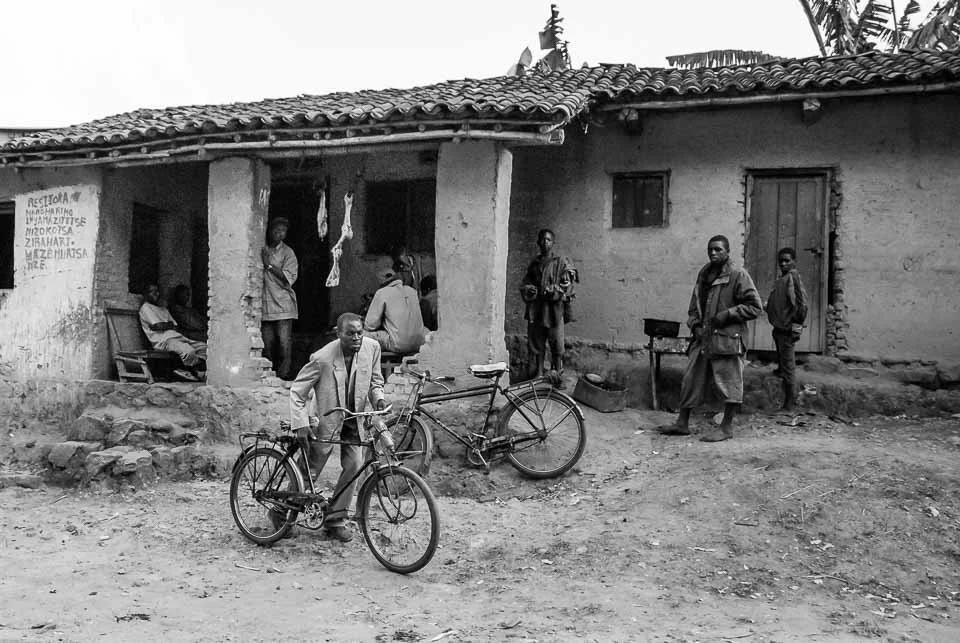 A black and white photo of a rural village scene with several people, a man with a bicycle, and a small building with a tiled roof.