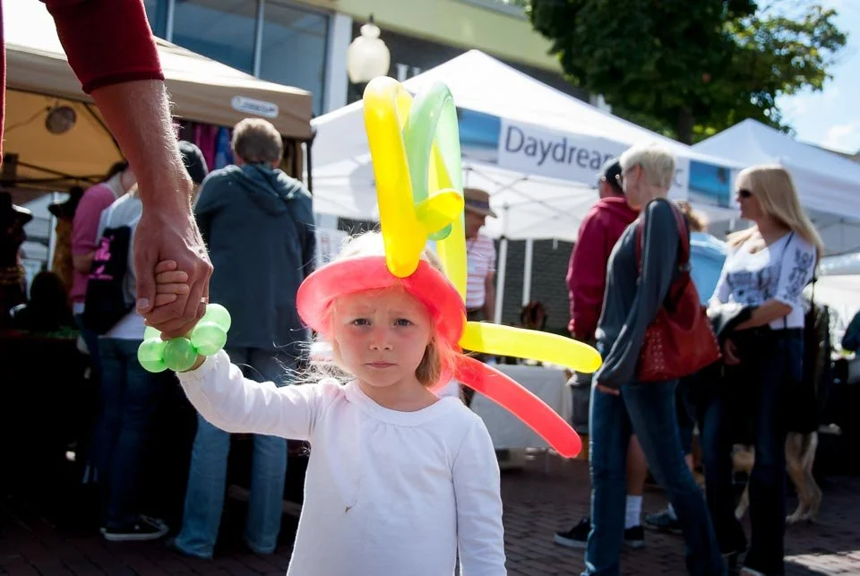 A young girl wearing balloon hat and holding hands with an adult at an outdoor market or fair with tents and people in the background.