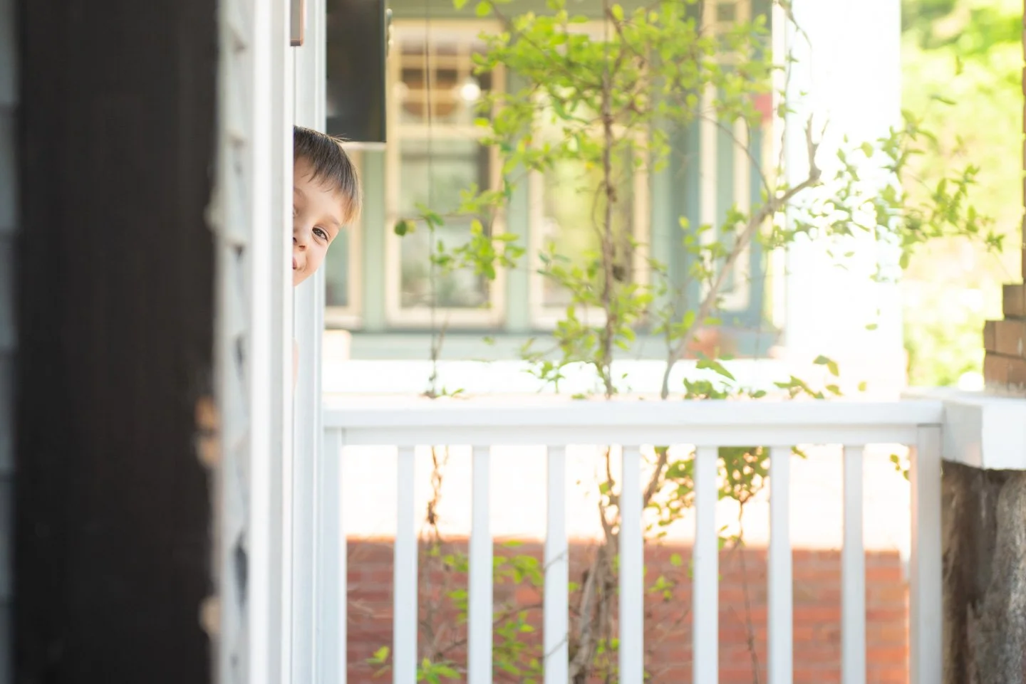A young boy peeking out from behind a vertical post on a porch, smiling.
