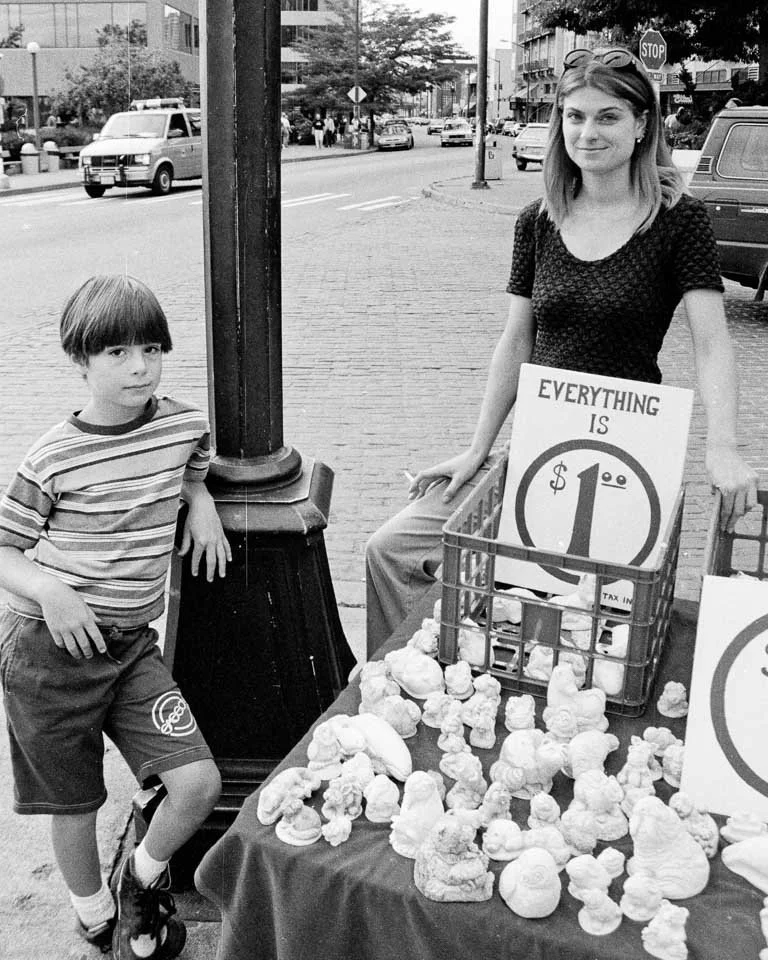 A woman selling carved soap figurines on a city sidewalk, with a boy leaning against a lamppost nearby. The woman has a sign indicating all items are priced at $1.00.
