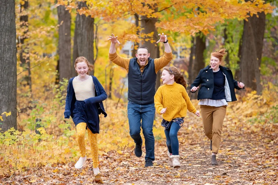 A family of four, two adults and two children, smiling and running along a leaf-covered trail in a park during autumn. The trees have vibrant orange, yellow, and green leaves.