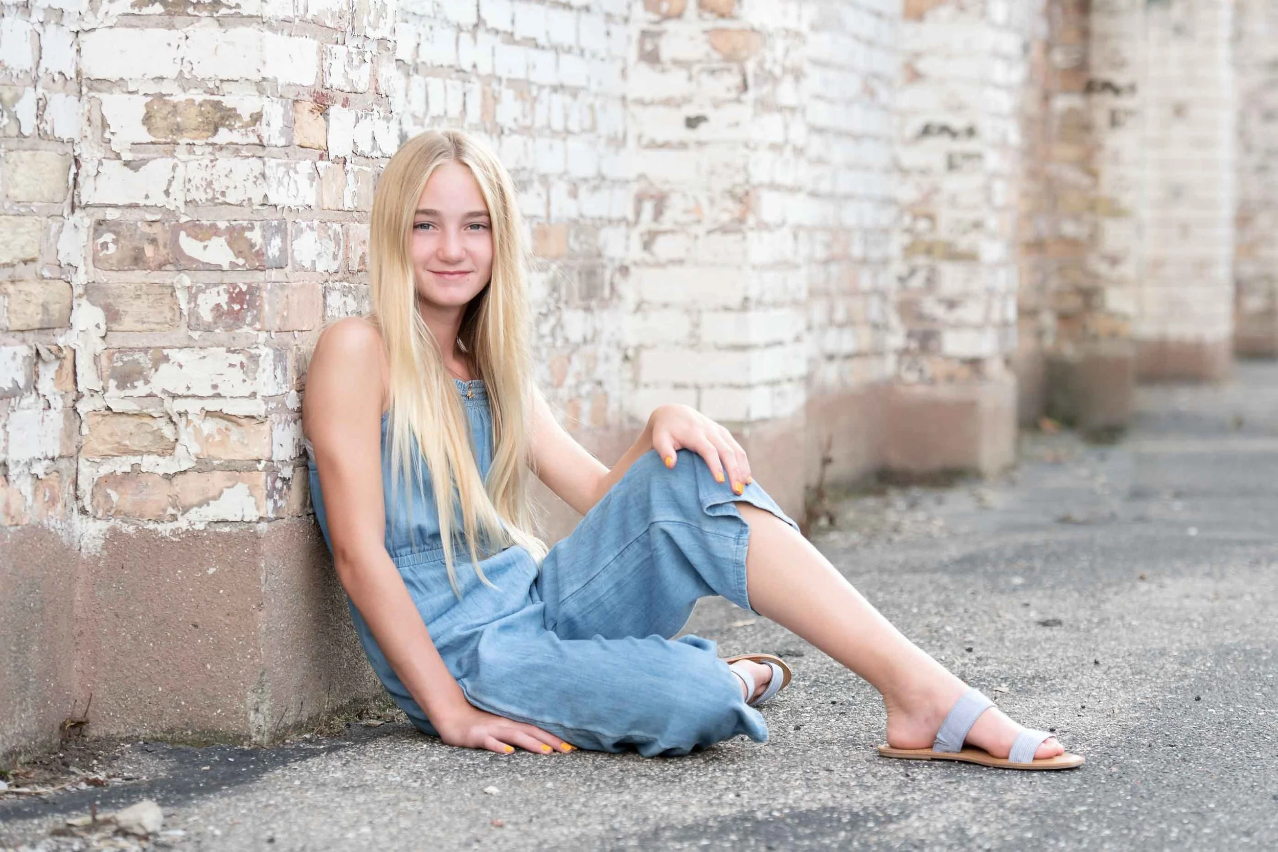 A young girl with long blonde hair sitting against a brick wall on a sidewalk, wearing a denim jumpsuit and sandals, looking at the camera with a slight smile.