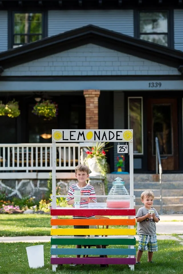 Two young boys selling lemonade from a colorful stand in front of a residential house with a porch, flowers, and hanging baskets.
