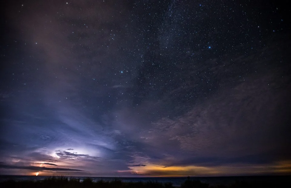 Night sky filled with stars over a landscape with some clouds and distant lights, possibly an ocean or open land.