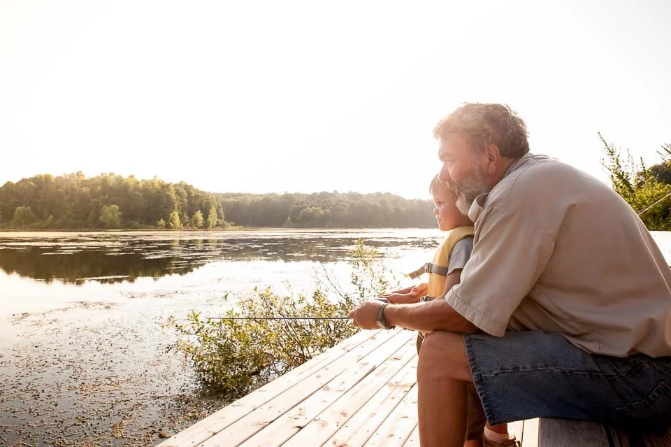 An older man and a young boy sit on a wooden dock fishing by a calm lake at sunset.