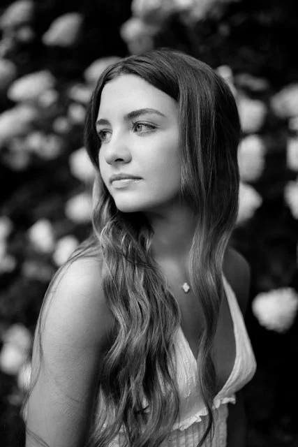 Black and white photo of a young woman with long, wavy hair, looking to her left, outdoors with blurred foliage in the background.