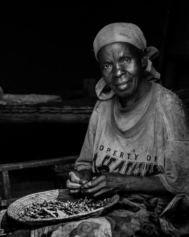 An elderly woman with a headscarf sitting and peeling peanuts or similar nuts from a woven basket.
