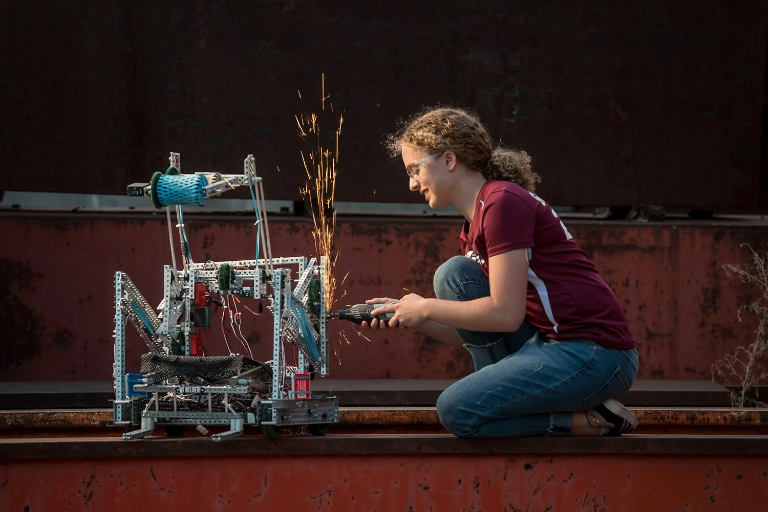 A girl with curly hair and glasses, wearing a maroon t-shirt and jeans, kneeling on the ground using a power tool to work on a robot made of metal and plastic parts on a rusty industrial surface.