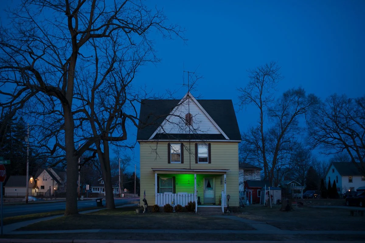 A yellow two-story house with a black roof, trees without leaves, and a porch with a green light during twilight.