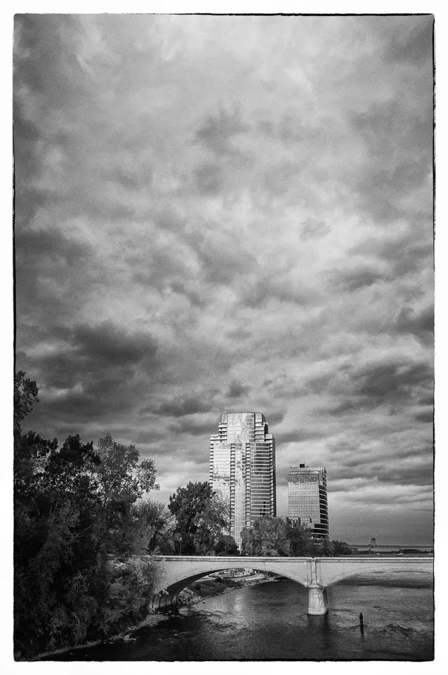Black and white photo of a city skyline with tall buildings over a river, with trees and a bridge in the foreground, and cloudy sky above.