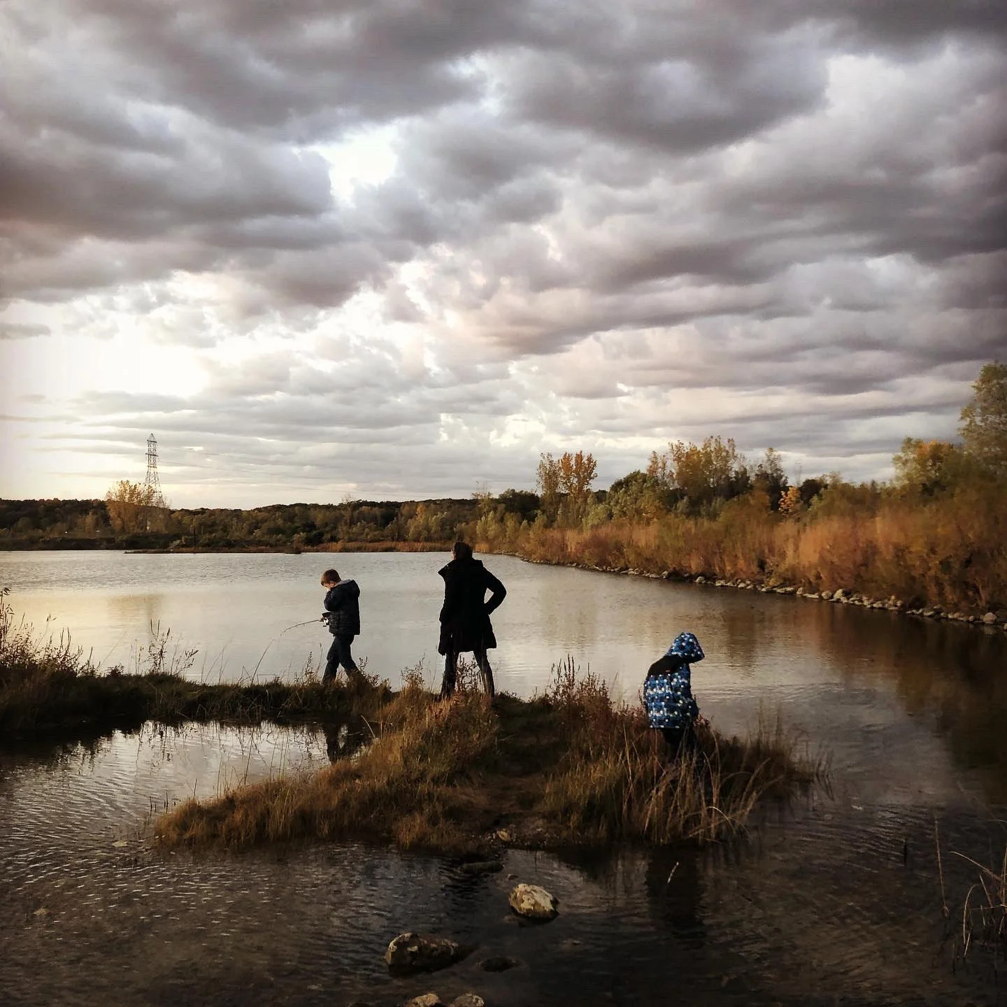 Three people fishing by a riverbank on a cloudy autumn day, with colorful trees in the background.