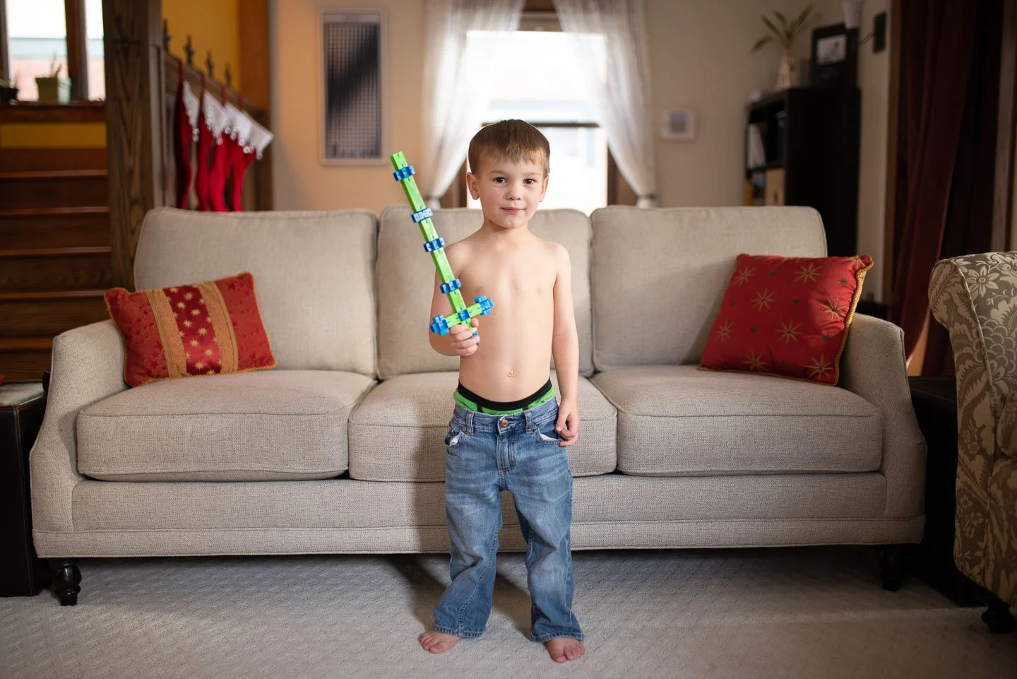 A young boy standing shirtless in front of a beige couch, holding a toy sword made of colorful building blocks, in a living room with red and gold pillows, curtains, and a staircase in the background.