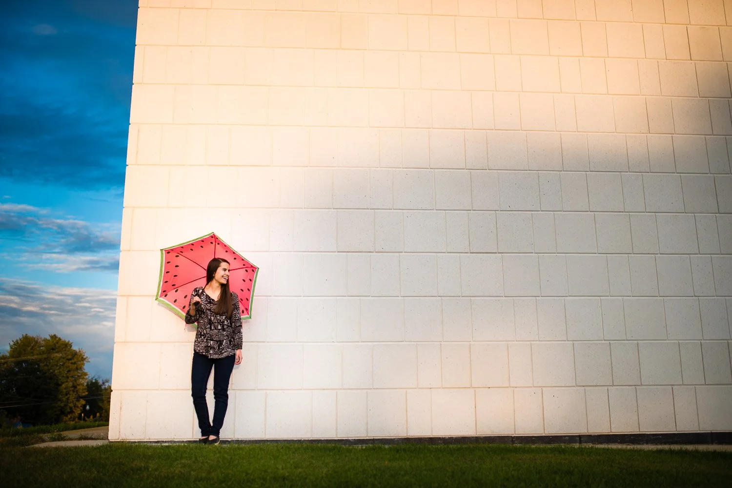 A woman standing outside near a large white wall, holding a pink watermelon-patterned umbrella, dressed in a features patterned top and black pants, smiling and looking to her left. The sky is partly cloudy with a mix of blue and white clouds, and th