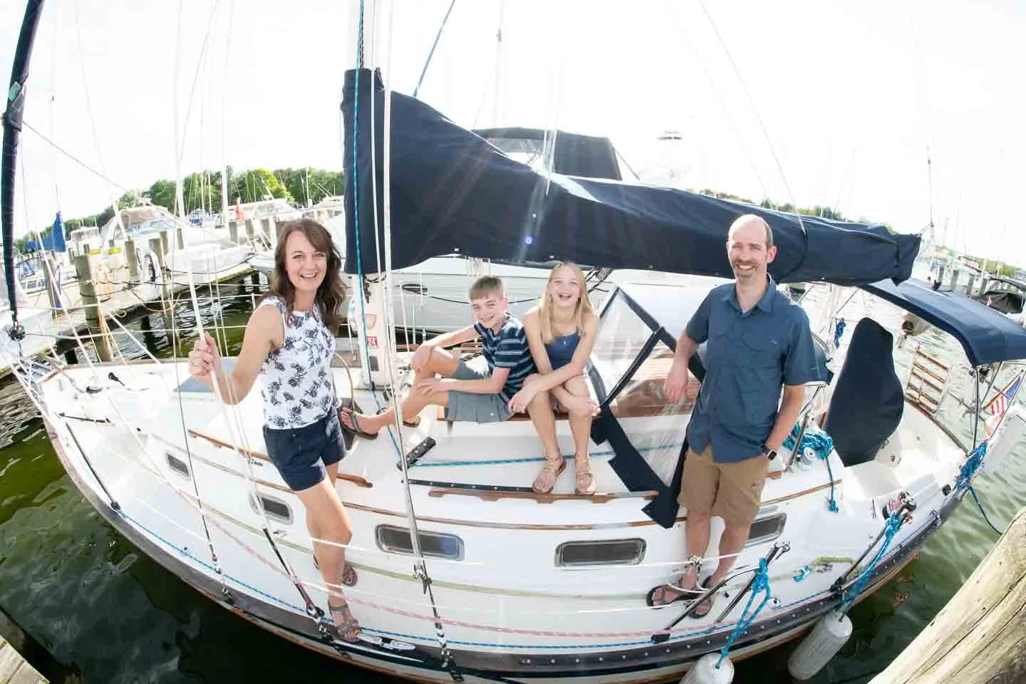 Family of four enjoying time on a sailboat docked at a marina, with 2 children sitting on the boat deck and two adults standing on the boat sides, all smiling.