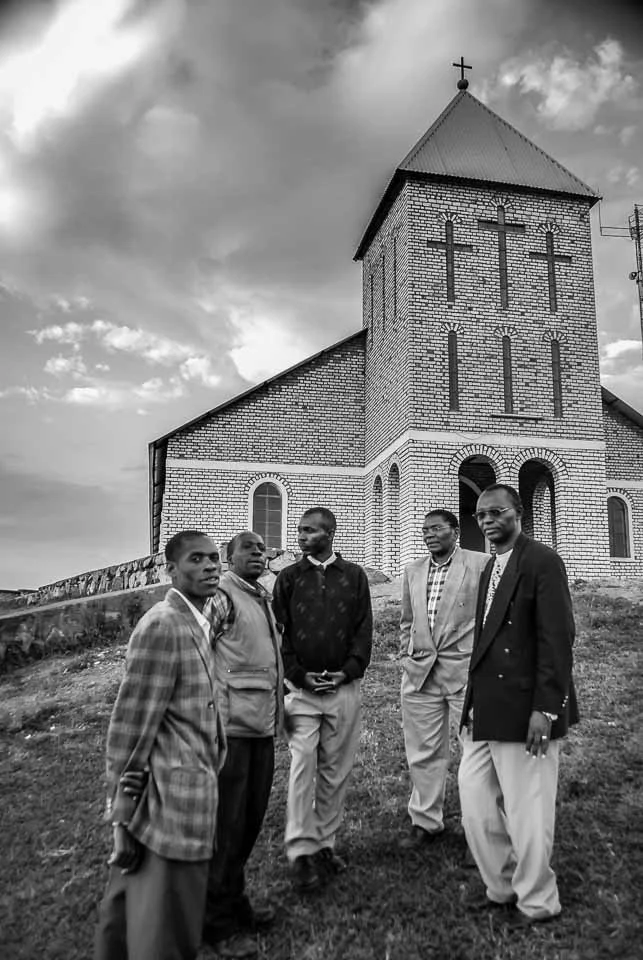 Five men stand on a grassy hill in front of a brick church with a steeple and cross, under a cloudy sky.
