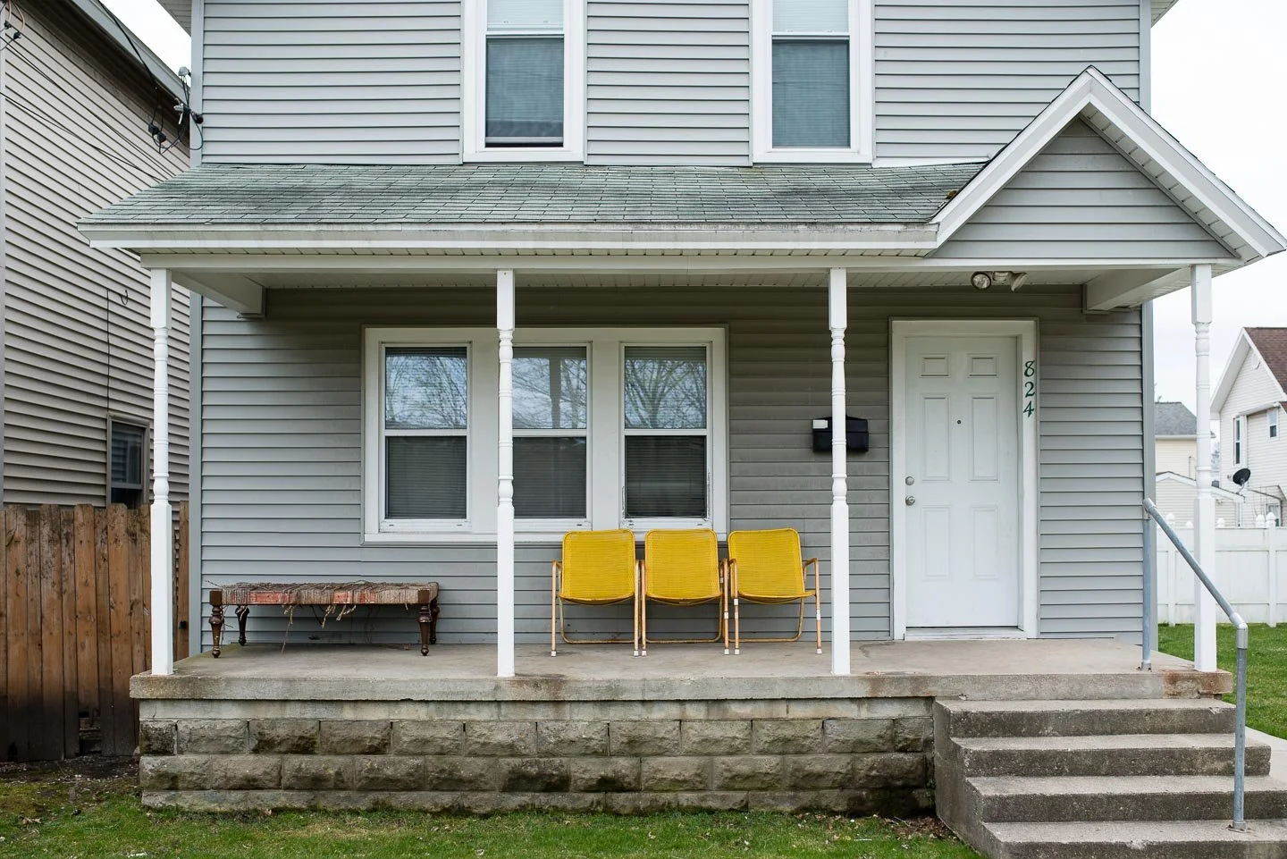 Front porch of a gray house with three yellow chairs, a small wooden bench, and a white door displaying the number 824. The porch has a concrete foundation, steps, and white columns supporting the roof.