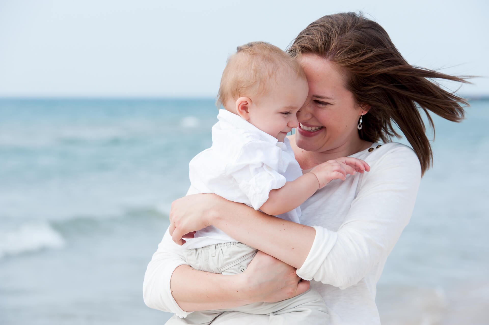 A woman and young boy at the beach, hugging and touching foreheads, with water and sky in the background.