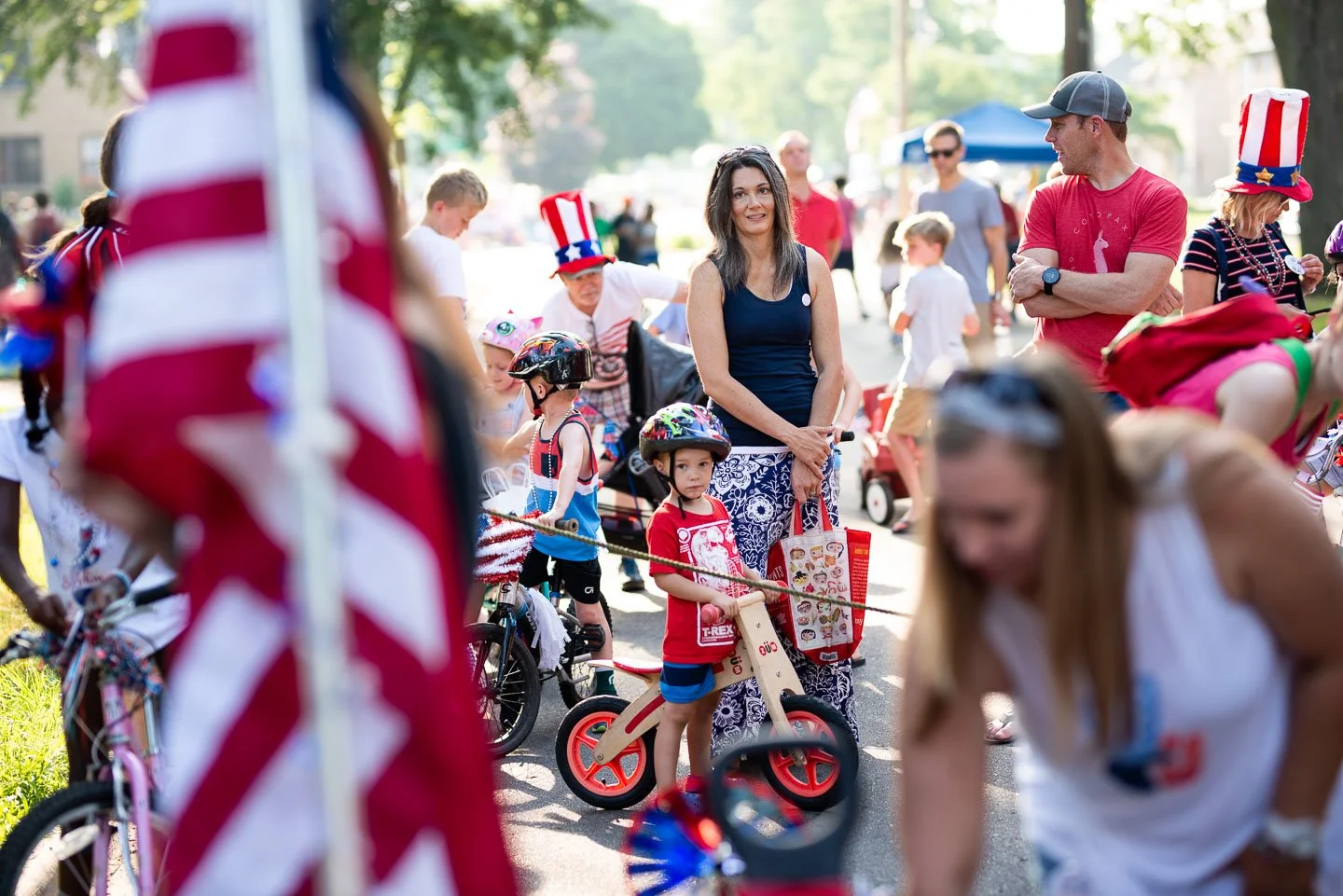 People celebrating at a 4th of July event, with children riding bikes and wearing patriotic accessories, and adults wearing red, white, and blue.