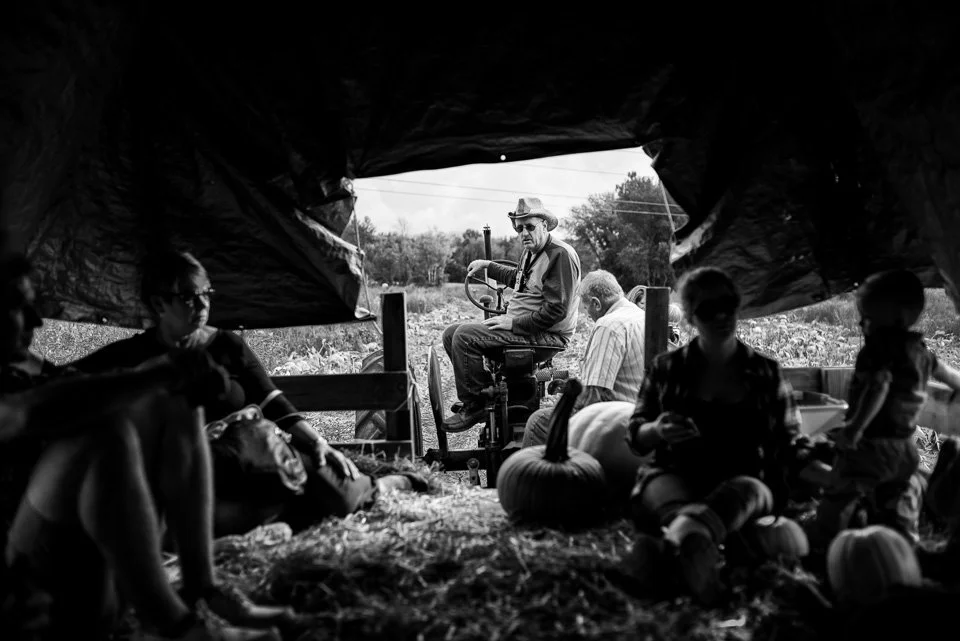 Scene of people in a barn, looking out toward a man on a tractor with pumpkins, in black and white.