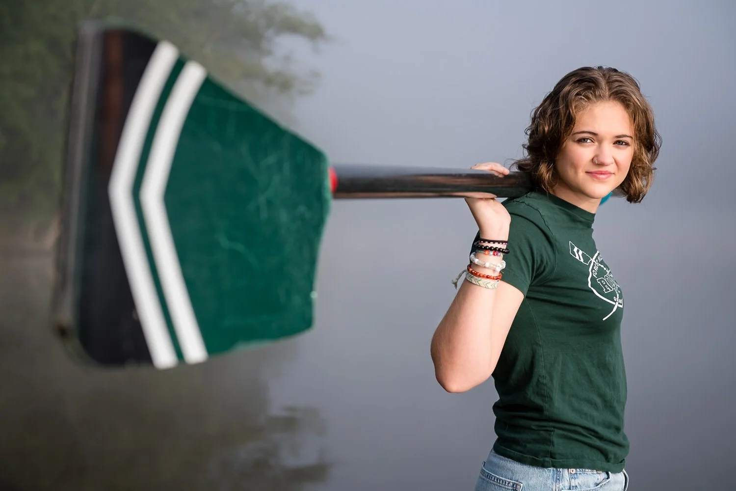 Young woman with short curly hair holding a green paddle over her shoulder near a body of water.