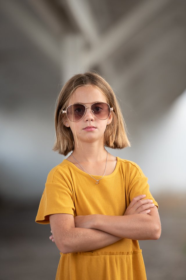 A young girl with short blonde hair wearing large sunglasses, a yellow dress, and a necklace, standing with arms crossed in an indoor setting.