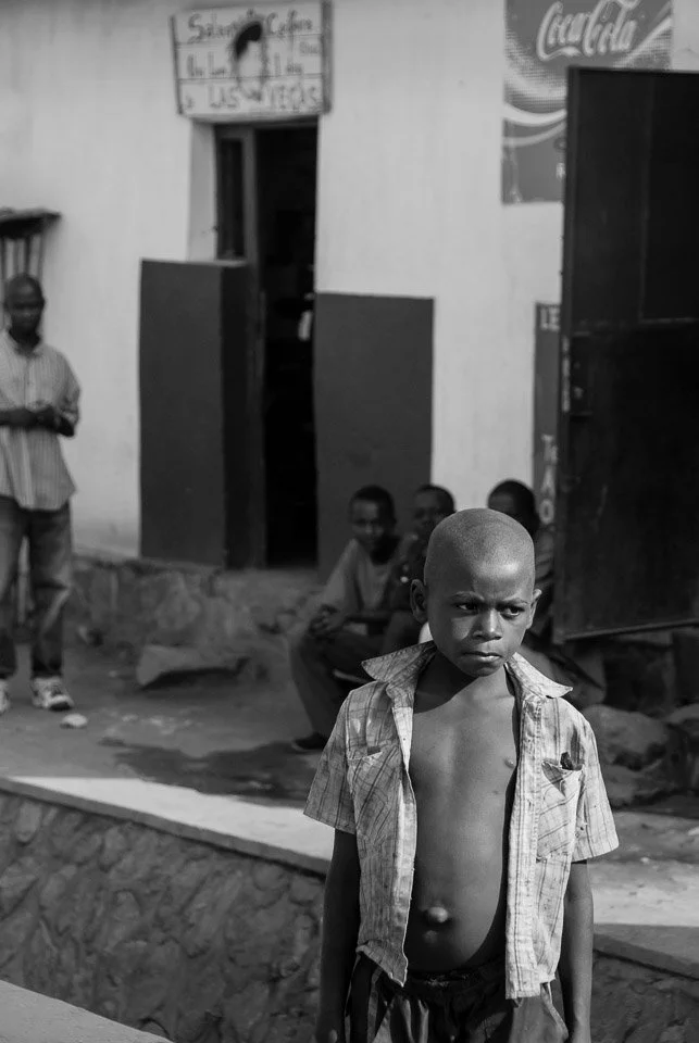 A group of children and a man standing outside a building. A young boy in the foreground has a serious expression, is shirtless with an unbuttoned shirt over his shoulders, and has a short haircut. In the background, three children are sitting, and a