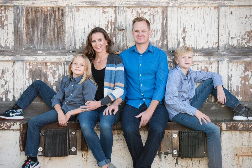 Family of four sitting on a weathered wooden wall, smiling at the camera. The mother is wearing a striped cardigan and jeans, sitting next to her teenage son, who is wearing a blue shirt and jeans. The young daughter has blonde hair and is wearing a 