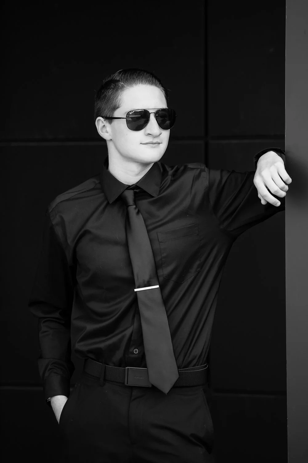 A young man in a black shirt, black tie, and sunglasses leaning against a dark wall.