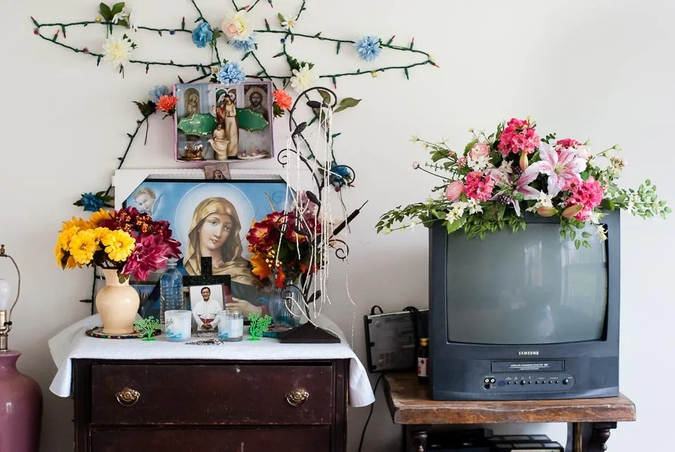 A home altar with religious icons, candles, and flowers, next to an old TV with a floral arrangement on top, against a white wall decorated with string lights and paper flowers.