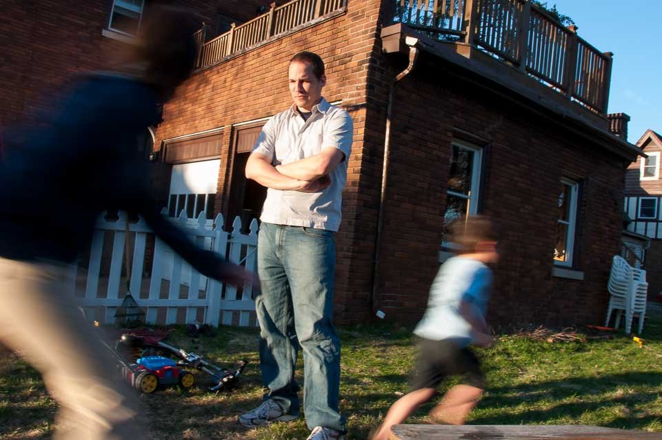 A man with crossed arms standing in a backyard with two children playing nearby, in front of a brick house with a white fence.