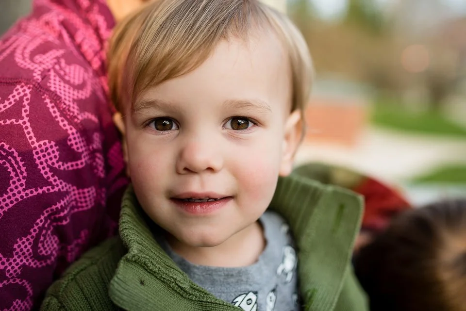 Close-up of a young boy with light brown hair and hazel eyes, smiling softly, wearing a green jacket and a gray T-shirt, outdoors with blurred background.